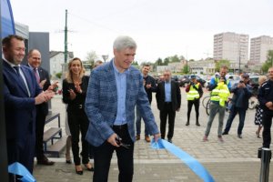 Burmistrz Miasta Czeladź Zbigniew Szaleniec przecina wstęgę na oficjalne otwarcie dworca w Czeladzi. Na drugim planie znajdują się osoby uczestniczące w otwarciu na świeżym powietrzu.