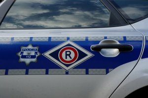 Zdjęcie przedstawia drzwi radiowozu policyjnego, na którym się znajduję logo z odznaką policyjną i napisem POLICJA 112 oraz emblemat oznaczający pojazd ruchu drogowego.