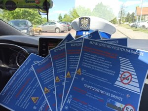 Zdjęcie przedstawia broszury informacyjne z najważniejszymi zmianami w przepisach ruchu drogowego, w tym regulacji związanych ze zmianami dotyczącymi bezpieczeństwa pieszych. Broszury znajdują się w radiowozie policyjnym, a za nimi czapka policyjna, szyba i różne pojazdy na drugim planie.