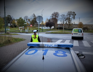 Na zdjęciu znajduje się policjantka przed radiowozem, który zaparkowany jest w okolicy przejścia dla pieszych.